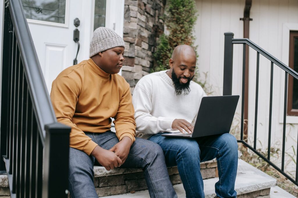 Positive bearded African American male remote worker sitting on stairs and typing on netbook keyboard with teen son